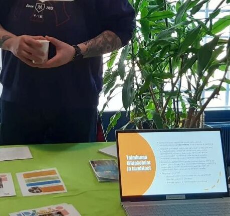 Henkilö seisomassa pöydän takana. Pöydällä Klubitalon esitteitä, sekä kannettava tietokone jossa pyörii Klubitalon toiminnasta kertova Powerpoint-esitys